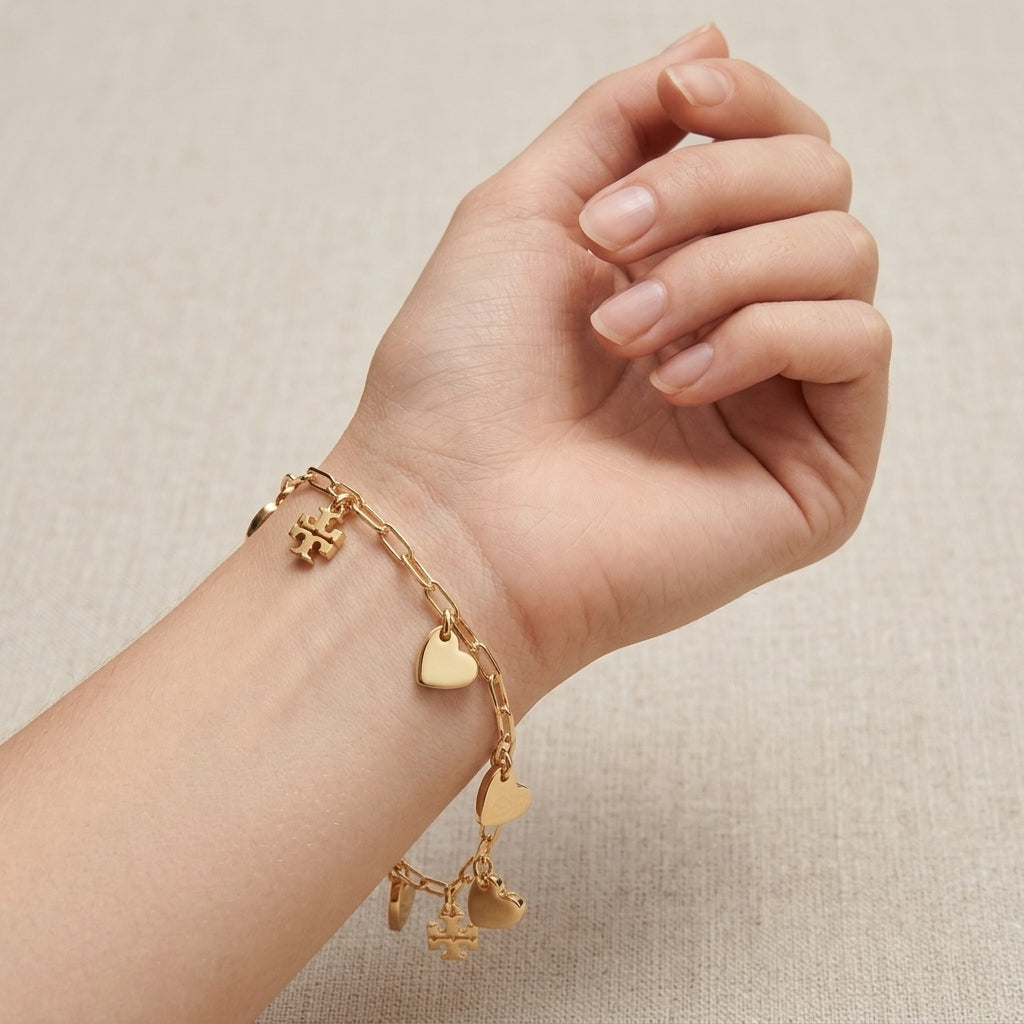 A gold charm bracelet with heart and cross charms, shown worn on a person's wrist against a neutral beige textured background.
