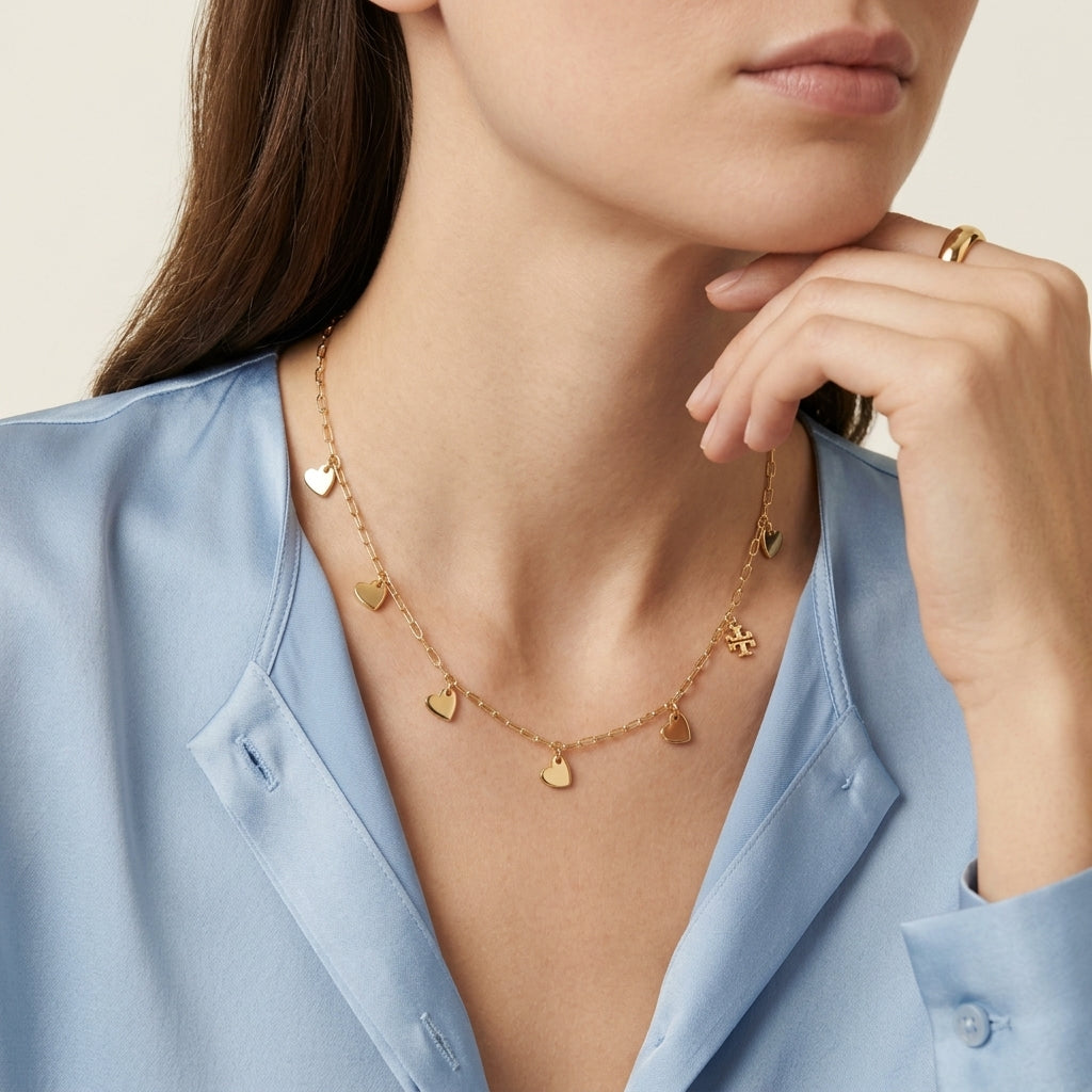 A close-up of a model wearing a delicate gold necklace with small heart and flower charms, paired with a light blue satin shirt.