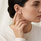 A close-up of a woman wearing multiple gold ear piercings and earrings, including bow-shaped studs, a heart-shaped stud, a pearl stud, and a star-shaped stud, along with a wide gold ring and a delicate gold necklace.