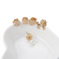 A set of five gold-tone stud earrings displayed on a white ceramic dish against a white background. The designs include a flower with clear crystals, a star, a heart, a small dog, and a single pearl stud.
