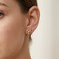 A close-up of a model's ear wearing a small gold stud earring shaped like a rocking horse, against a soft off-white background.