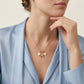 A close-up lifestyle shot of a model wearing a delicate gold necklace with a charm featuring a small white pearl and a gold heart, styled with a light blue satin blazer.