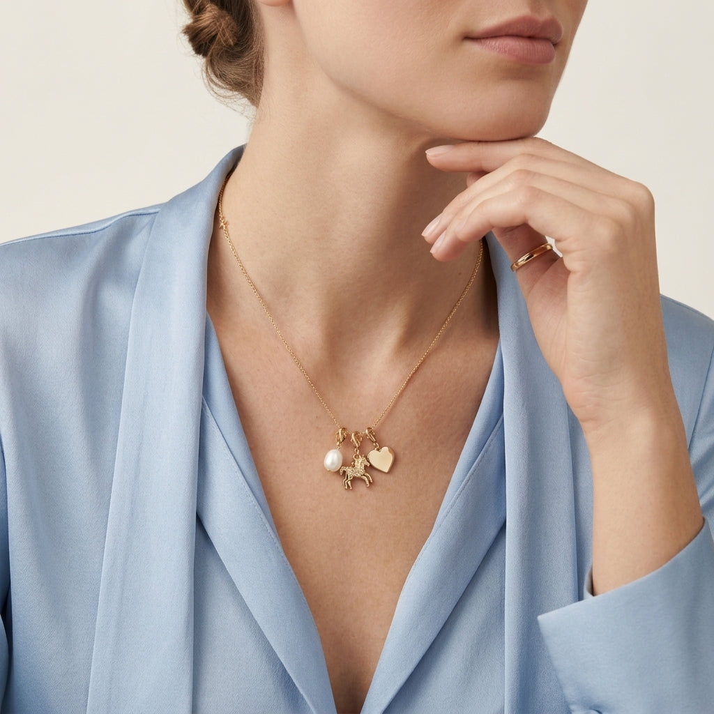 A close-up lifestyle shot of a model wearing a delicate gold necklace with a charm featuring a small white pearl and a gold heart, styled with a light blue satin blazer.