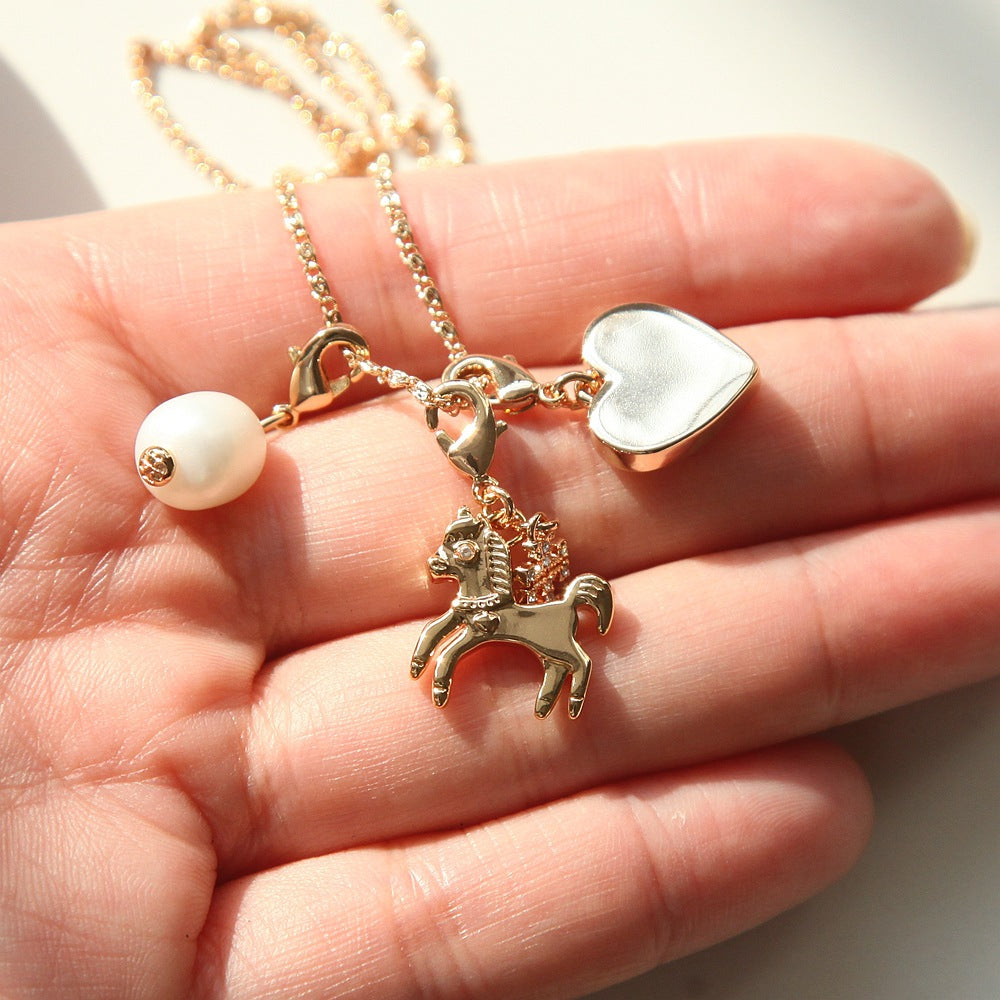 A gold-tone charm necklace with a horse, heart, and pearl bead, held in a person's hand.