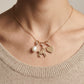 A close-up of a person's neck and upper chest wearing a delicate gold necklace with three charms: a white pearl, a small gold horse figure, and a flat gold heart. The necklace is styled with a beige knit sweater.