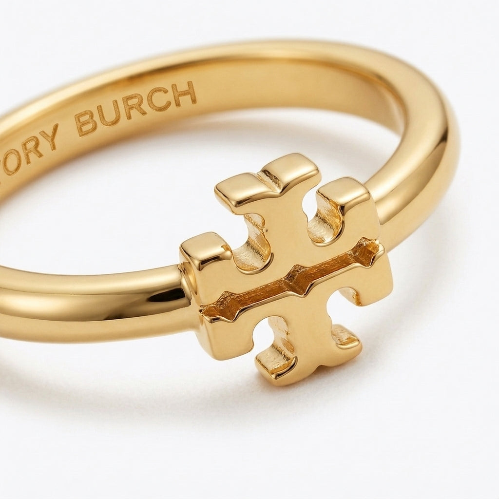 A gold ring featuring a polished interlocking cross logo charm, with the brand name 'TORY BURCH' engraved on the inner band, set against a clean white background.