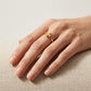 A delicate gold ring with a small sculpted floral or starburst motif worn on a person's ring finger, resting on a textured beige fabric surface.