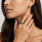 A close-up lifestyle shot of a model wearing a gold signet ring with a green enamel background and a gold monogram logo, alongside a matching gold earring.