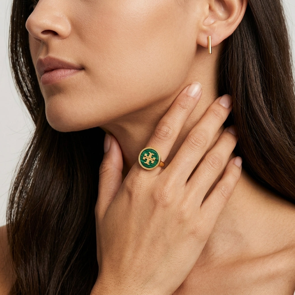 A close-up lifestyle shot of a model wearing a gold signet ring with a green enamel background and a gold monogram logo, alongside a matching gold earring.