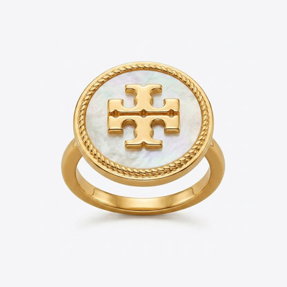 A gold ring featuring a round white mother-of-pearl inlay with a raised gold monogram logo in the center, set against a light gray background.