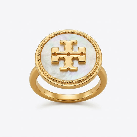 A gold ring featuring a round white mother-of-pearl inlay with a raised gold monogram logo in the center, set against a light gray background.
