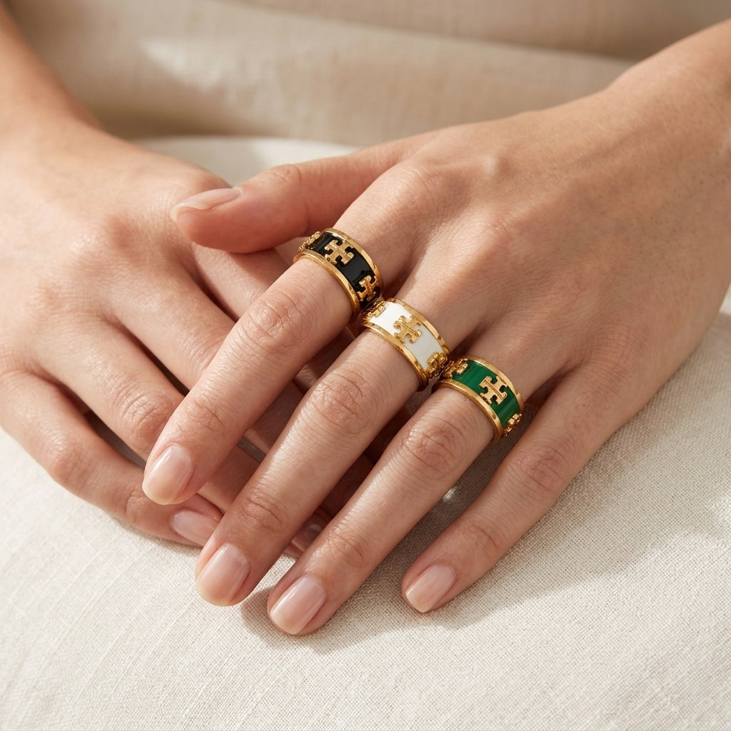 Three gold rings with colorful enamel inserts are shown on a person's fingers. One ring has black enamel, one has white enamel, and one has green enamel, all with gold monogram-style details.