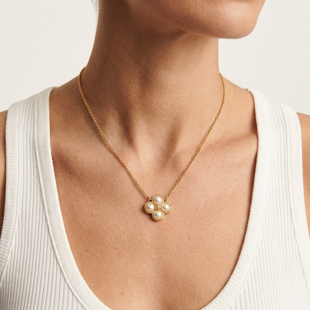 A close-up of a model wearing a delicate gold chain necklace with a small flower-like pendant made of five white pearls, styled with a white ribbed tank top.