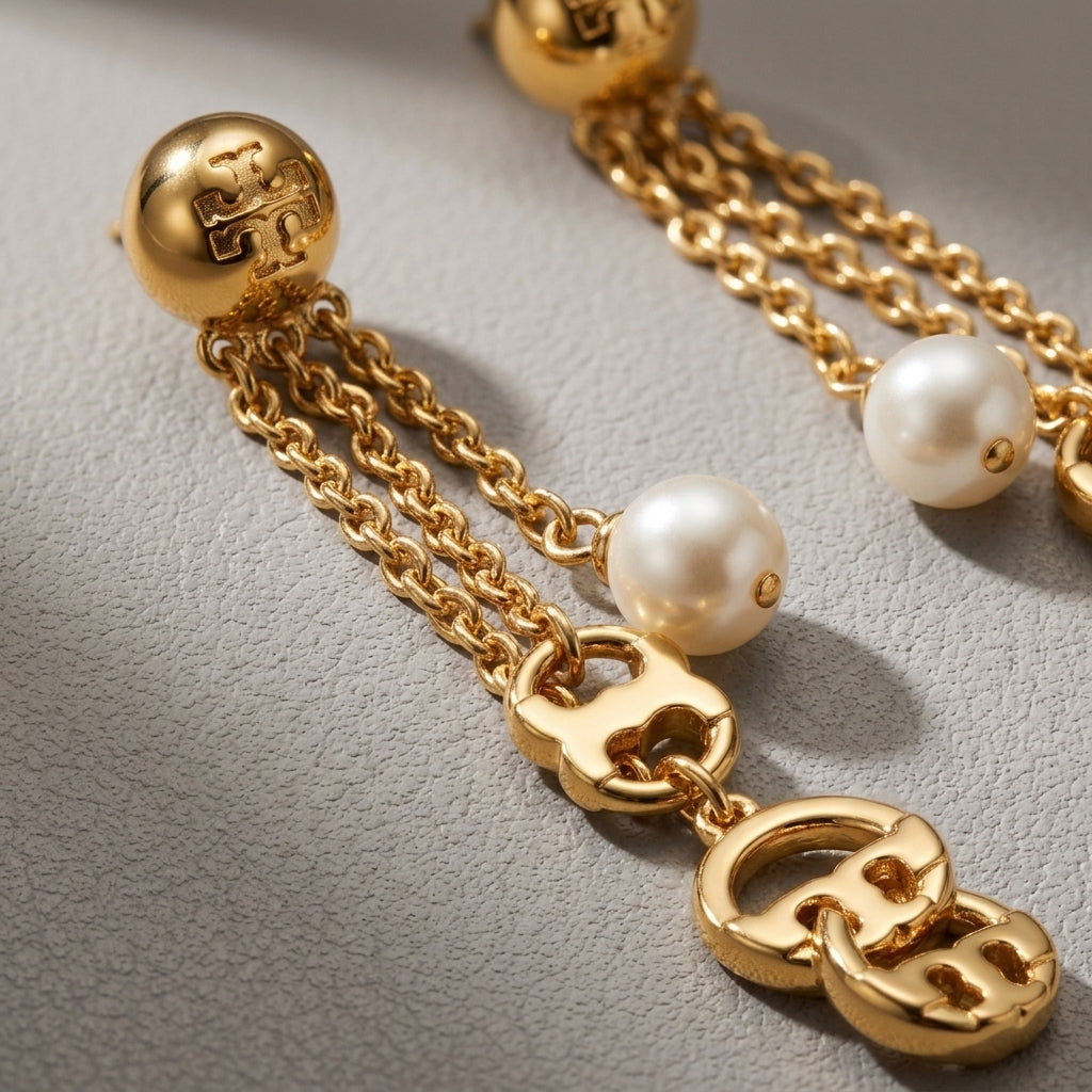 Gold-tone dangle earrings with pearl beads, chain links, and interlocking logo charms on a textured light gray surface.