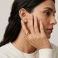 Woman wearing gold earrings and ring, with hand near face on neutral background