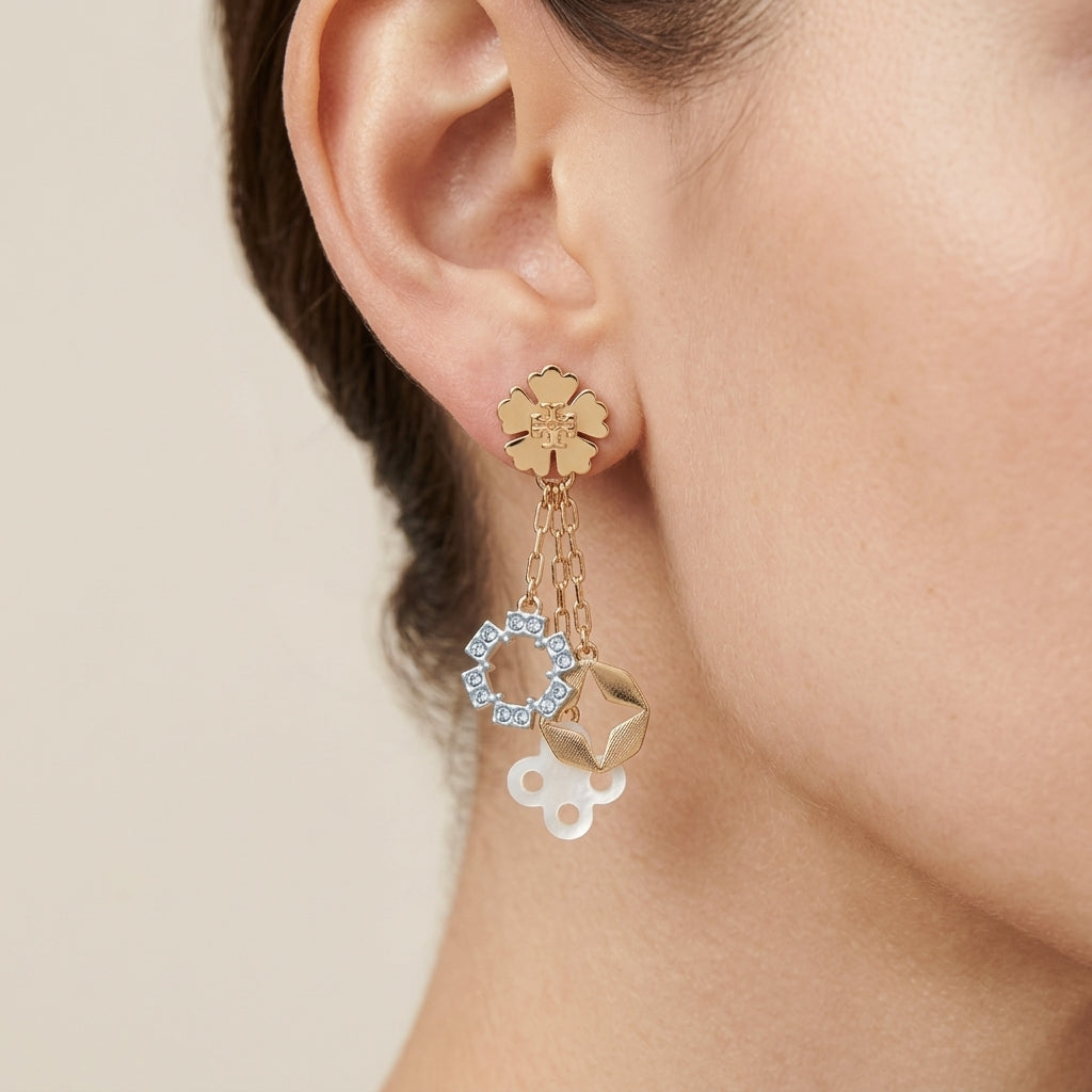 A close-up of a model's ear wearing a gold flower stud earring with dangling charms, including a clear crystal ring, a gold geometric charm, and a white cloud charm, against a soft beige background.