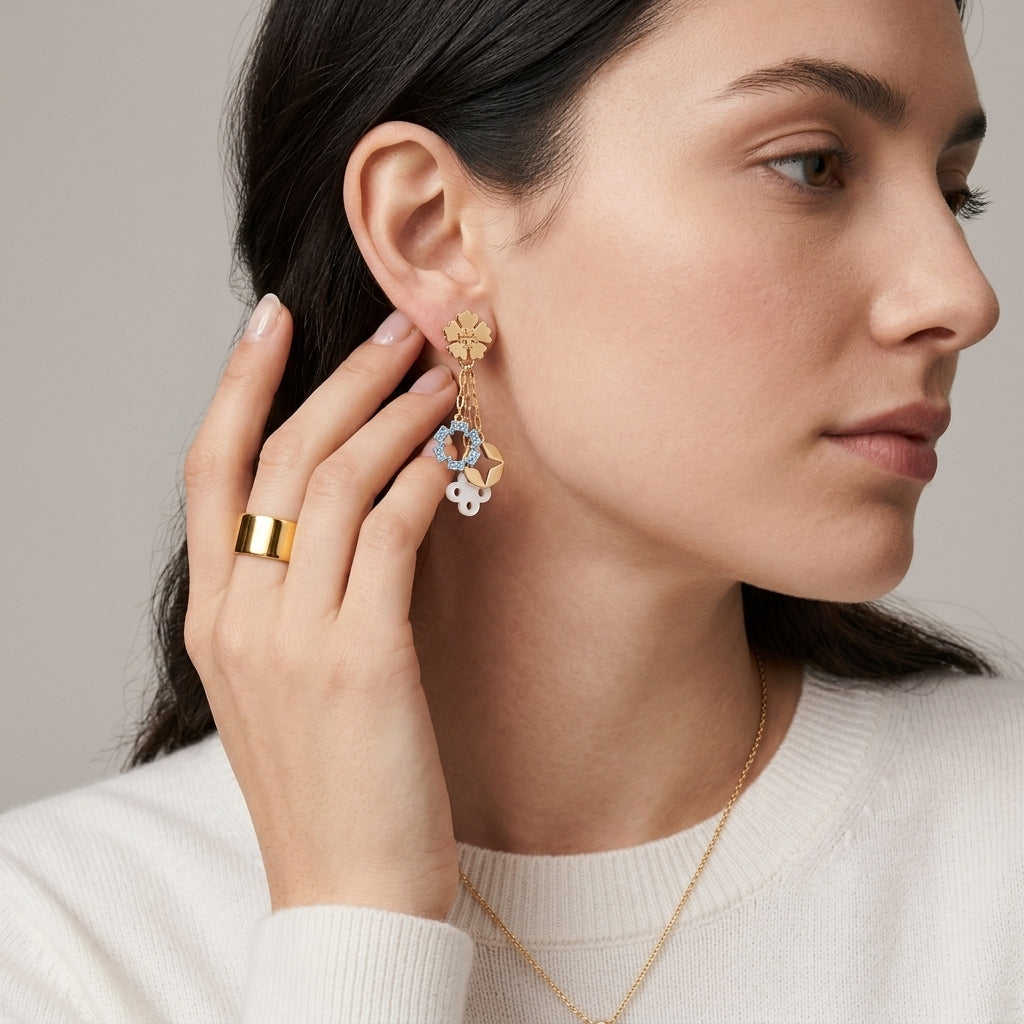 A close-up of a woman wearing a gold dangle earring with floral and butterfly details, along with a matching gold ring and necklace, against a neutral beige background.
