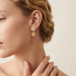 A close-up of a woman's ear wearing a delicate gold dangle earring with a white enamel four-petal stud and a dangling gold flower charm, set against a soft beige background.
