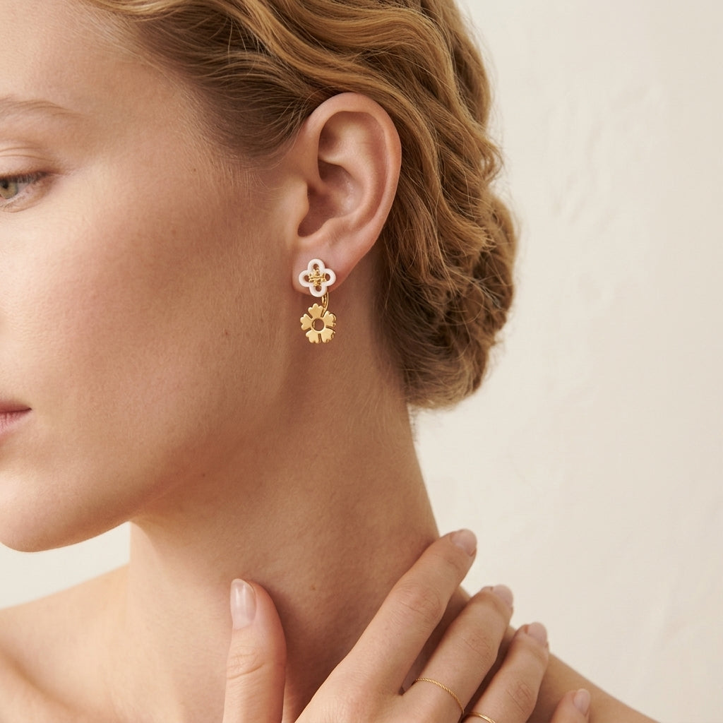 A close-up of a woman's ear wearing a delicate gold dangle earring with a white enamel four-petal stud and a dangling gold flower charm, set against a soft beige background.