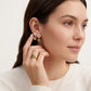 A close-up of a woman wearing gold floral stud earrings and a matching gold ring, shown against a clean light background.