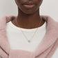 A close-up of a model wearing a delicate gold chain necklace with a small flower-shaped pendant, styled over a white t-shirt and a dusty pink knit cardigan.