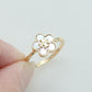 A gold ring with a four-petal flower face, featuring a white inlay and a small gold monogram in the center, held between fingers against a light gray background.