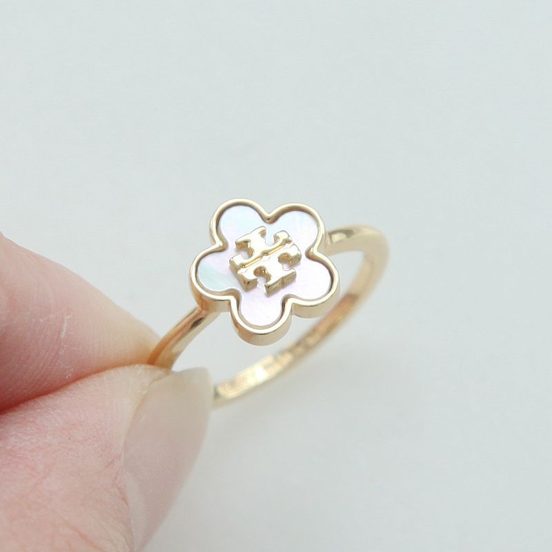 A gold ring with a four-petal flower face, featuring a white inlay and a small gold monogram in the center, held between fingers against a light gray background.