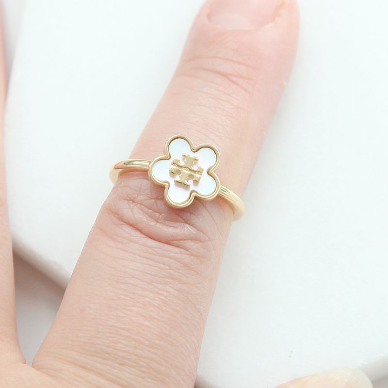 A gold ring with a white enamel four-petal flower face, worn on a finger against a clean white background.