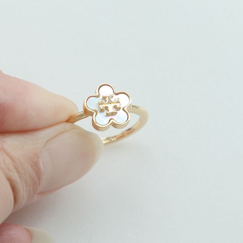 A gold ring shaped like a small flower with a white inlay, held between two fingers against a plain light background.