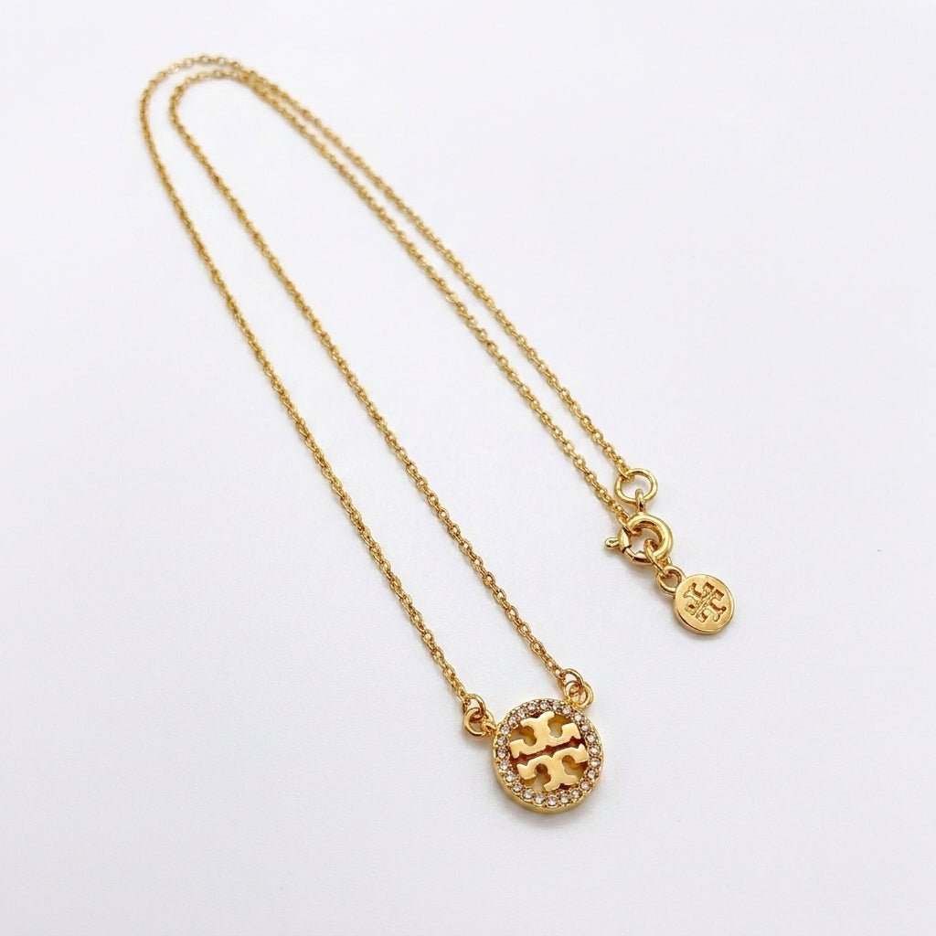 A gold chain necklace featuring a circular pendant with a gold 'T' logo surrounded by small clear crystals, and a small round tag charm, displayed on a clean white background.