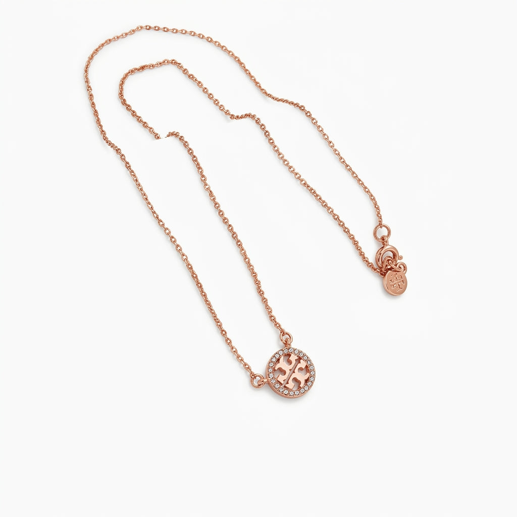 A delicate rose gold chain necklace featuring a circular pendant with a raised 'T' monogram and a small matching charm, displayed on a clean white background.