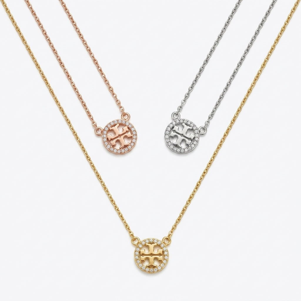 Three circular pendant necklaces on chains, shown on a light gray background. The necklaces are in yellow gold, rose gold, and silver tones, each featuring a pavé-set circular logo pendant with a central letter design.