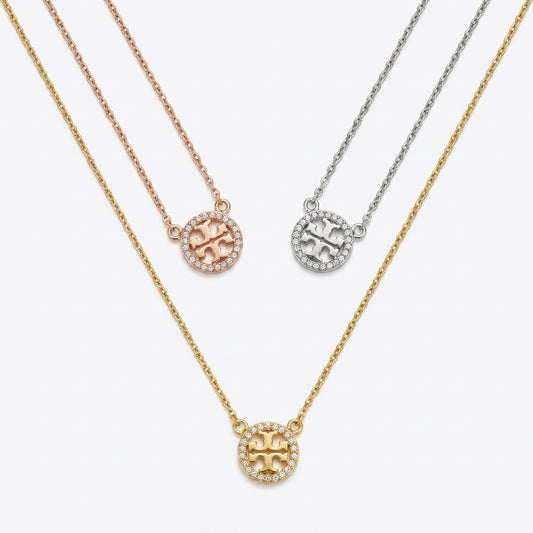 Three circular pendant necklaces on chains, shown on a light gray background. The necklaces are in yellow gold, rose gold, and silver tones, each featuring a pavé-set circular logo pendant with a central letter design.