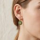 A close-up of a model's ear wearing a gold hexagonal dangle earring with green enamel and a gold monogram logo, against a light textured wall.