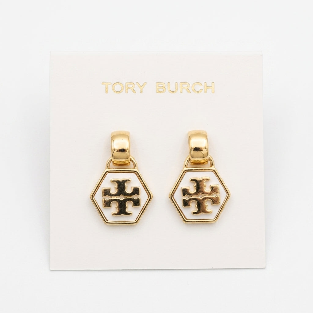 A pair of gold-tone hexagonal dangle earrings with a white enamel center and a black Tory Burch cross logo, displayed on a white backing card printed with the brand name.