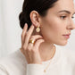 A close-up of a woman wearing a delicate gold dangle earring with a sunflower center charm, along with a matching gold ring and necklace, set against a soft white background.