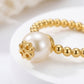 A gold beaded bracelet featuring a large white pearl and a decorative gold flower clasp, displayed on a white surface.