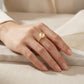 A close-up of a hand wearing a delicate gold ring with a round white pearl, resting on a light beige fabric surface.