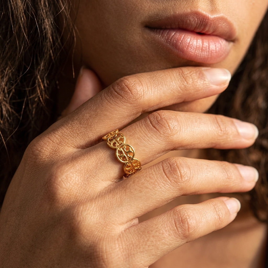 A close-up of a person's hand wearing a delicate gold ring with interlocking circular motifs, set against a soft-focus background of dark hair and skin tones.