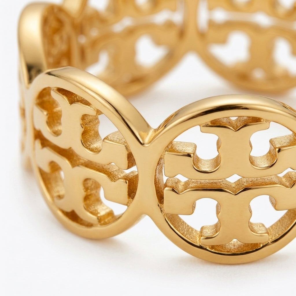 A gold-tone metal ring with an openwork interlocking monogram pattern, shown in a close-up studio shot on a white background.