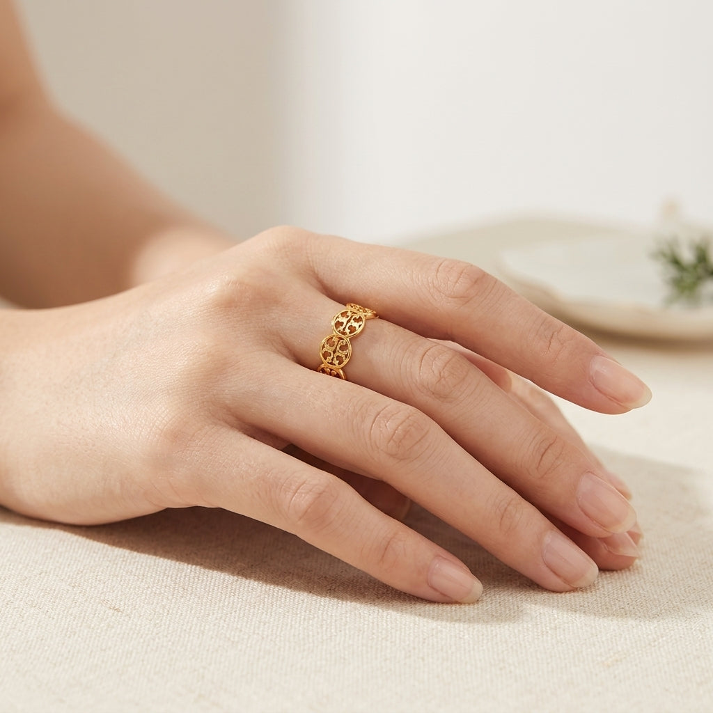 A delicate gold ring with an intricate openwork geometric pattern, worn on a woman's ring finger. The hand is resting on a textured beige fabric surface with a soft-focus background.