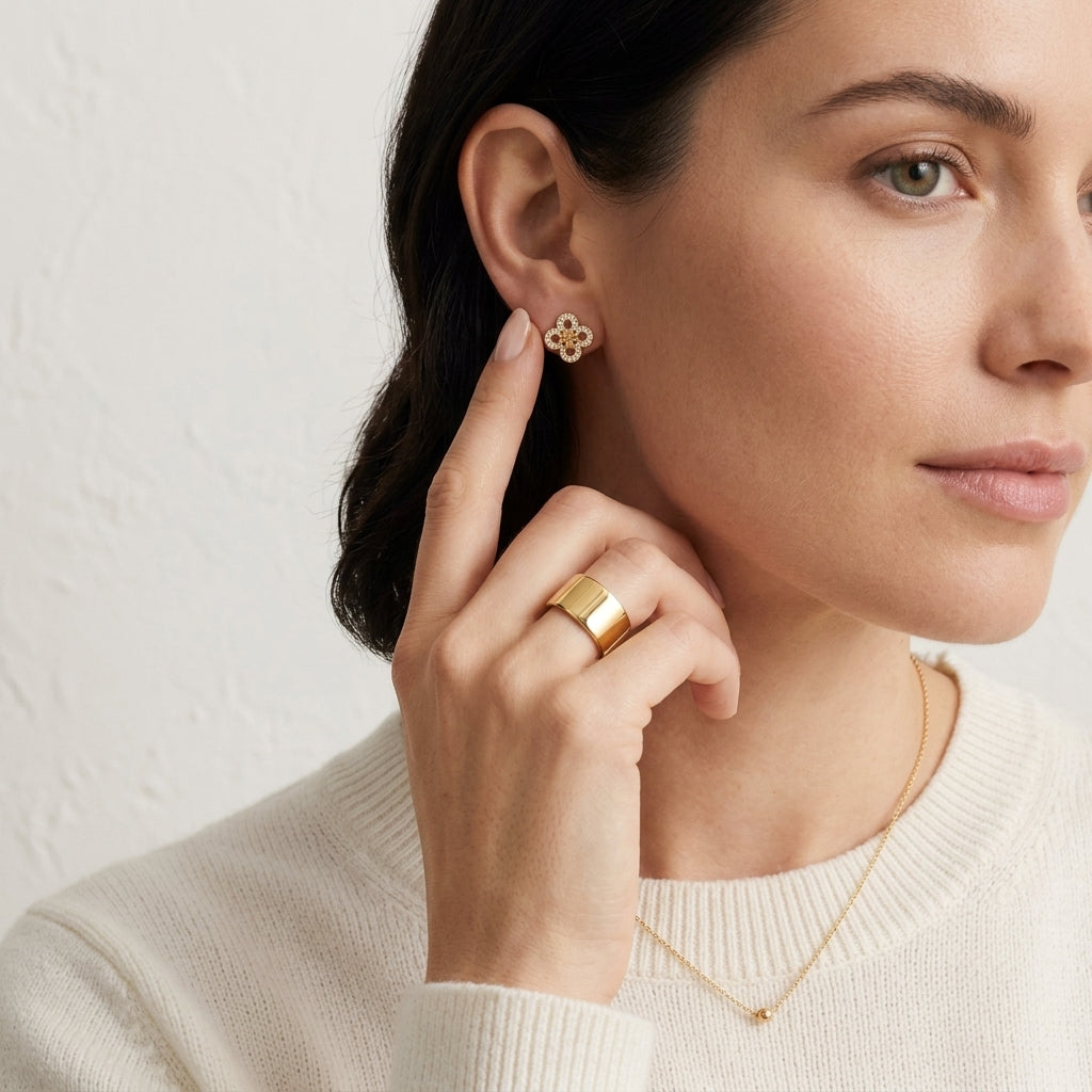 A close-up lifestyle shot of a woman wearing a delicate gold four-petal flower stud earring and a matching gold ring, set against a soft, textured off-white background.