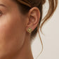 A close-up of a woman's ear wearing a small gold four-petal flower stud earring with tiny red and green gemstone accents, against a light neutral background.
