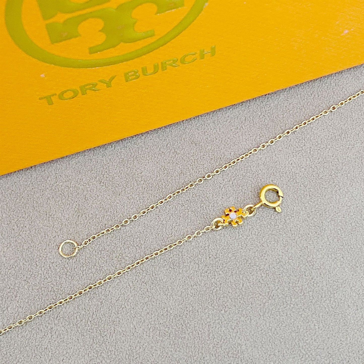 Gold necklace with a clasp on a gray surface with Tory Burch branding in the background.