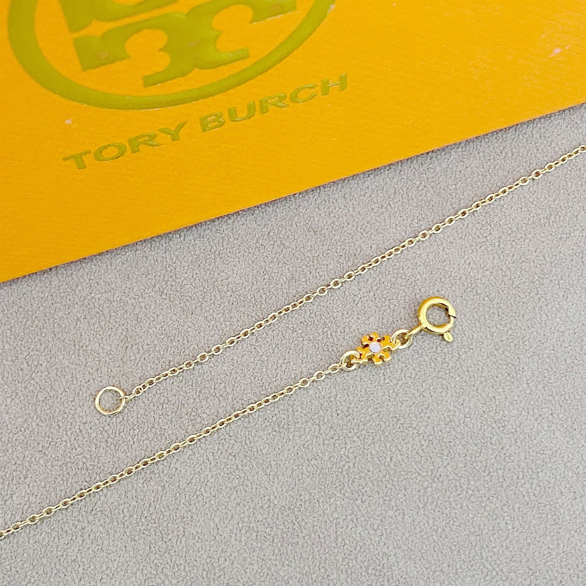 Gold necklace with a clasp on a gray surface with Tory Burch branding in the background.