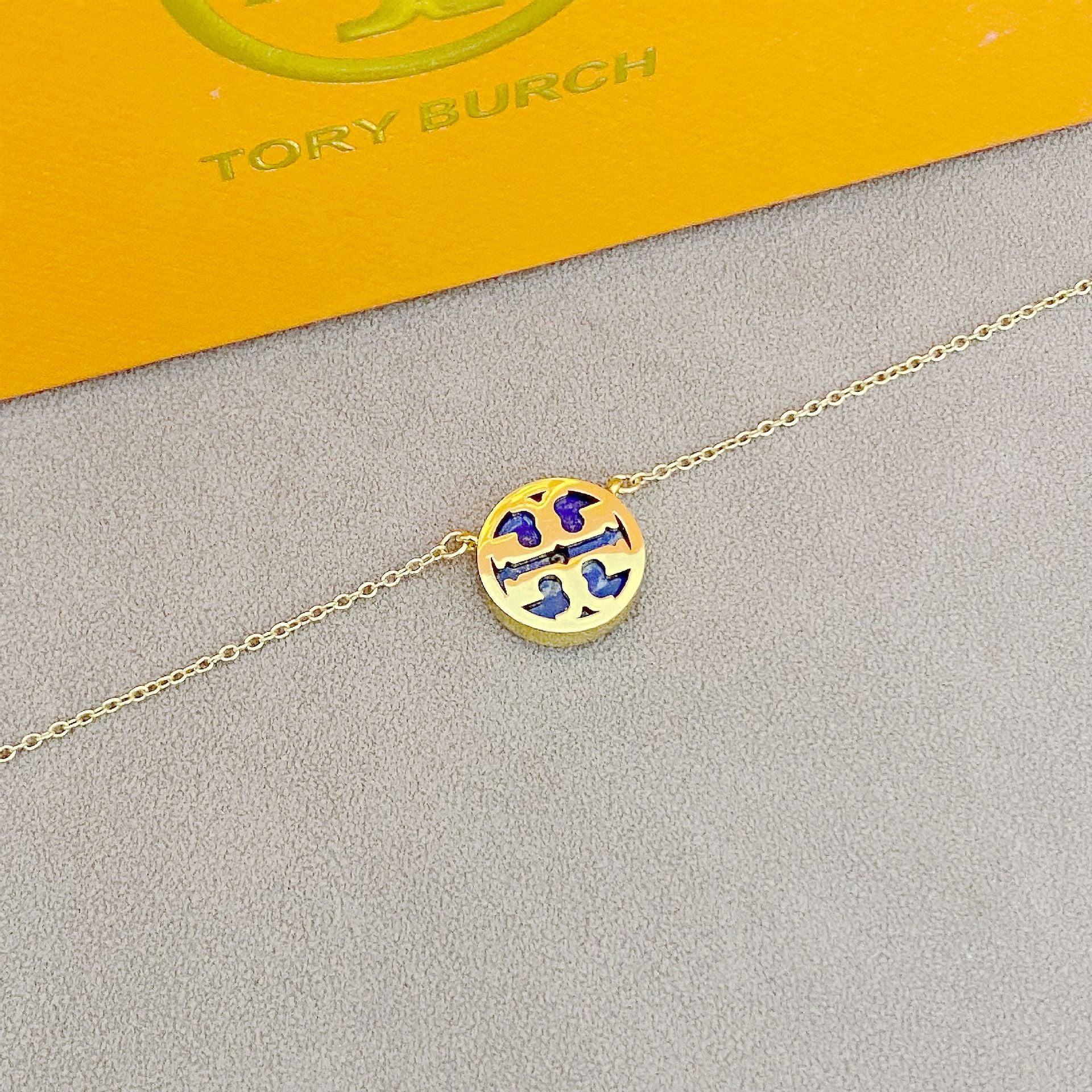 Gold necklace with a round pendant on a gray surface, with 'Tory Burch' branding in the background.