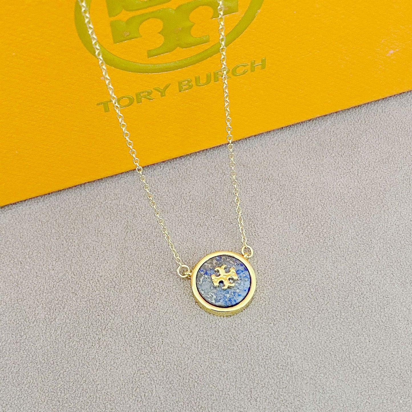 Necklace with a round pendant on a gray surface with a Tory Burch box in the background.