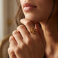 A close-up lifestyle shot of a model wearing a gold ring with a stylized, openwork geometric or floral-like design on her finger.