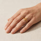 A gold ring with a textured, organic floral or honeycomb-like top, shown worn on a person's ring finger against a neutral beige fabric background.