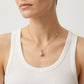 A close-up of a model wearing a delicate gold chain necklace with a small gold disc charm and a single pearl pendant, styled with a white ribbed tank top against a light gray background.